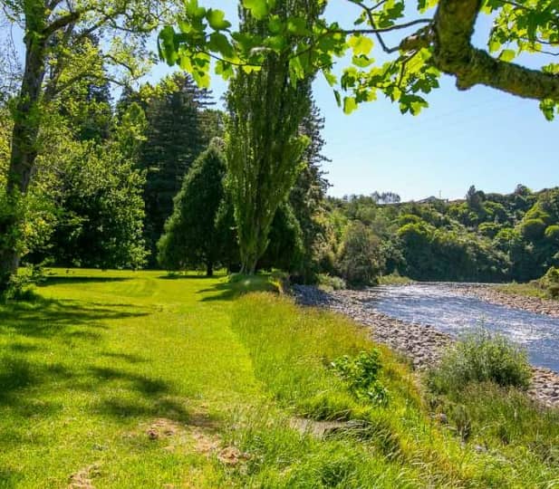 A wide view of a lush green riverside lawn at Tūpare Gardens, bordering a rocky river with tall trees and rolling hills under a bright blue sky. Credit: Leisure Time Travel