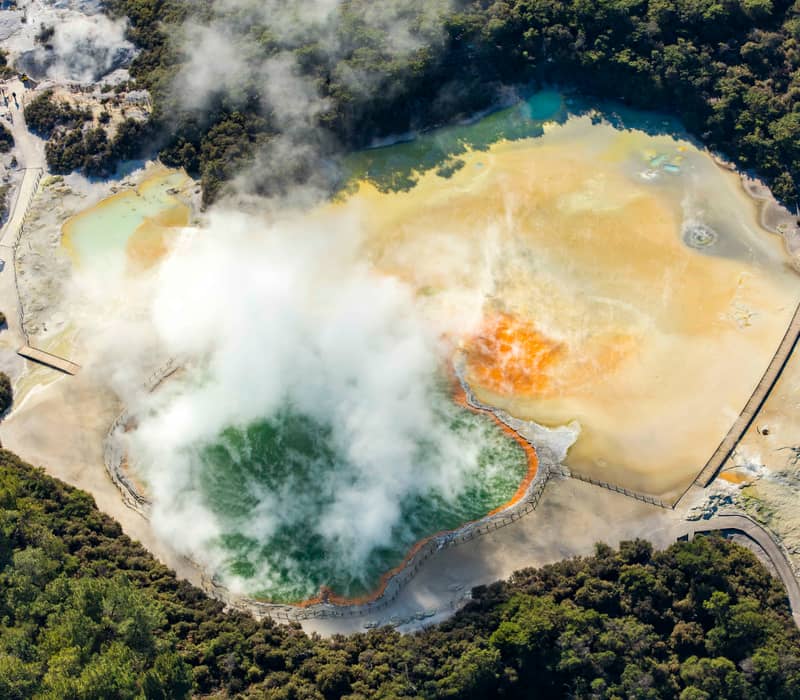 Aerial shot of a colorful geothermal hot spring emitting steam with forest and boardwalks nearby Credit: New Zealand Fine Touring