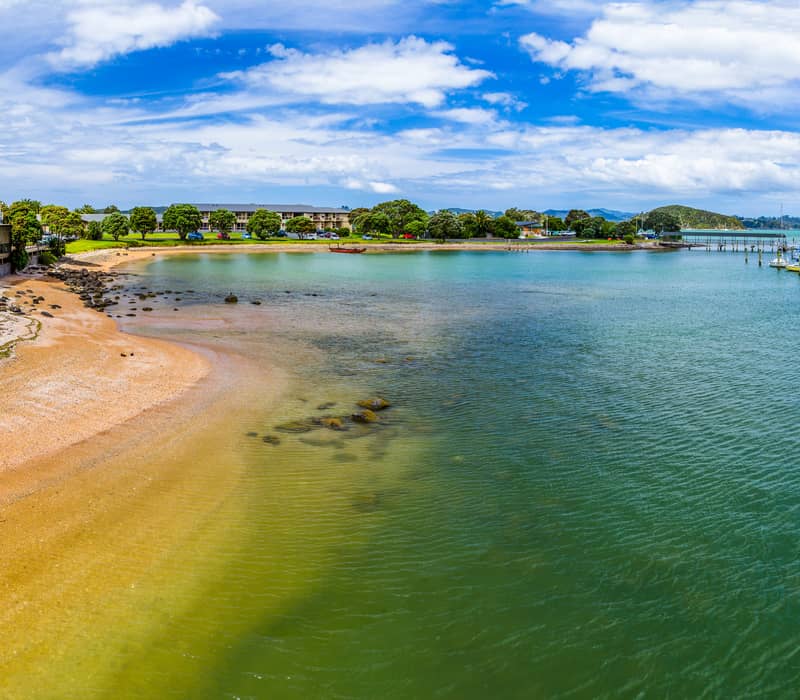 A panoramic view of Te Ti Bay in Paihia, showing a golden sand beach, clear green-blue water with moored boats, and beachfront buildings under a bright blue sky. Credit: Fine Tours Group - photographer unknown