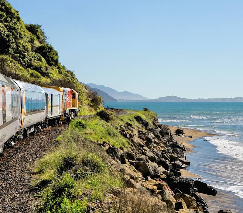 Train traveling along a coastal railway with the ocean and beach nearby under a clear sky. Credit: Great Journeys
