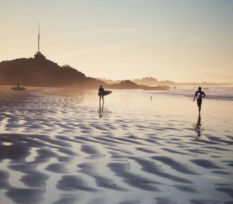 Surfers walking on a beach at sunset carrying surfboards with rippled sand and waves in the background. Credit: Great Journeys