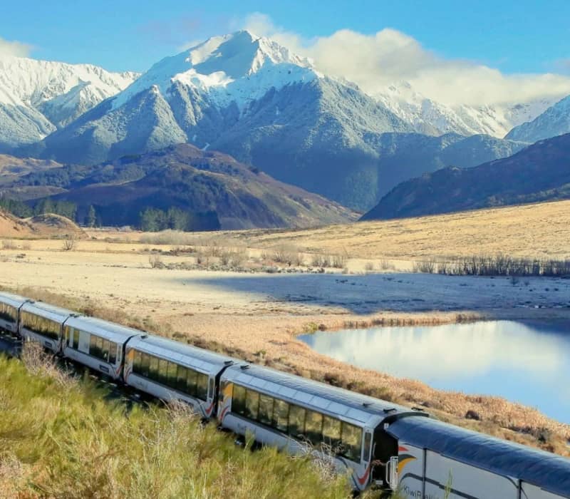 Train traveling through a mountain valley with snow-covered peaks and a calm lake. Credit: Great Journeys