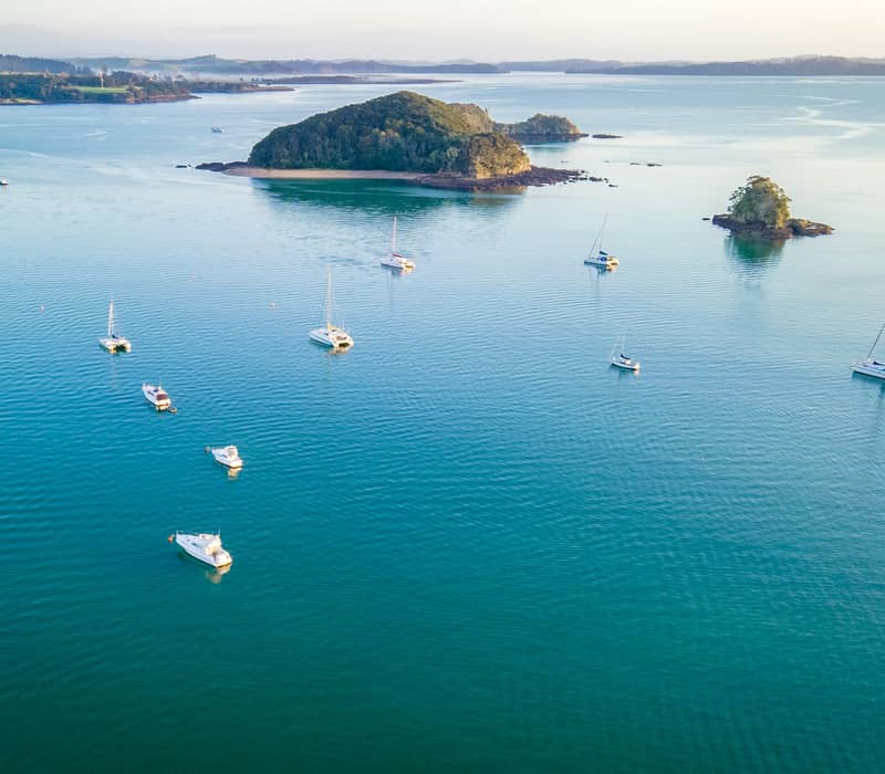 An aerial high-angle view of multiple white yachts, catamarans, and leisure boats anchored on calm, shimmering turquoise waters in a bay with lush green islands in the distance under a bright sky. Credit: Unsplash