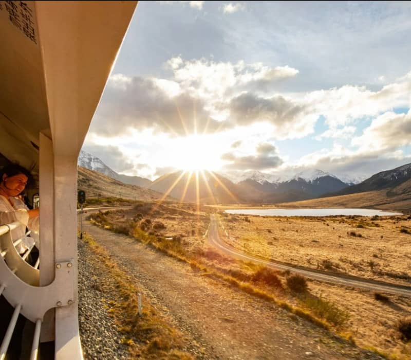 People on a train looking at a sunset over mountains and a lake in a wide valley. Credit: Great Journeys