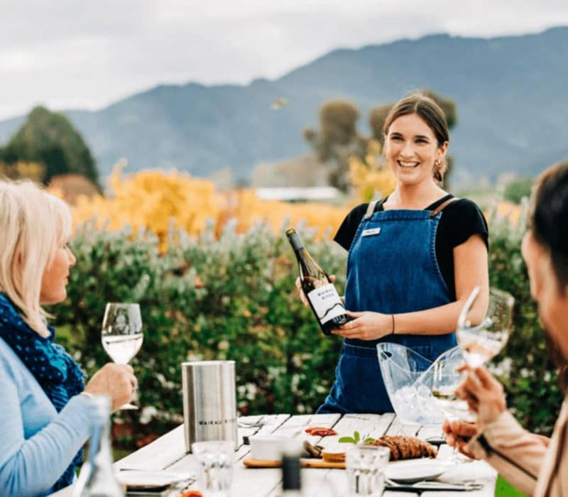 People enjoying outdoor wine tasting with a smiling host pouring wine at a vineyard with mountains behind. Credit: Great Journeys