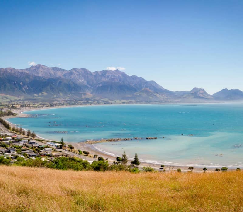 Coastal bay with clear blue water, town by the shore, mountains in background under a blue sky. Credit: Great Journeys
