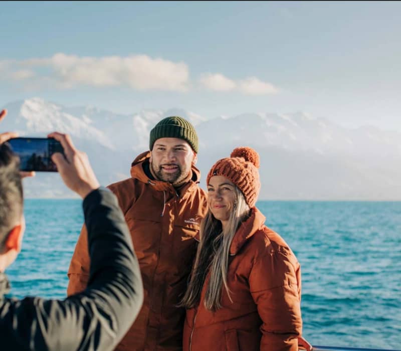 Couple in warm jackets and beanies posing by a lake with snowy mountains behind. Credit: Great Journeys