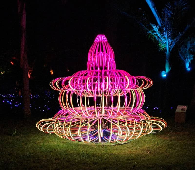 A vibrant neon light sculpture in the shape of a tiered fountain or pagoda, glowing with pink and orange lights in a dark park setting at night. Credit: Leisure Time Travel