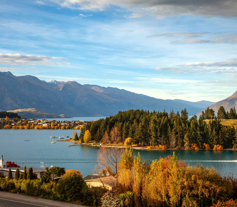Beautiful view of Queenstown Bay and the forested Queenstown Gardens peninsula surrounded by autumn foliage and mountains under a blue sky. Credit: Ling Hua - Unsplash