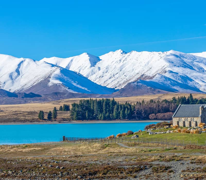 Stone church by bright blue lake with snow-capped mountains in background under clear sky. Credit: Pure Discoveries