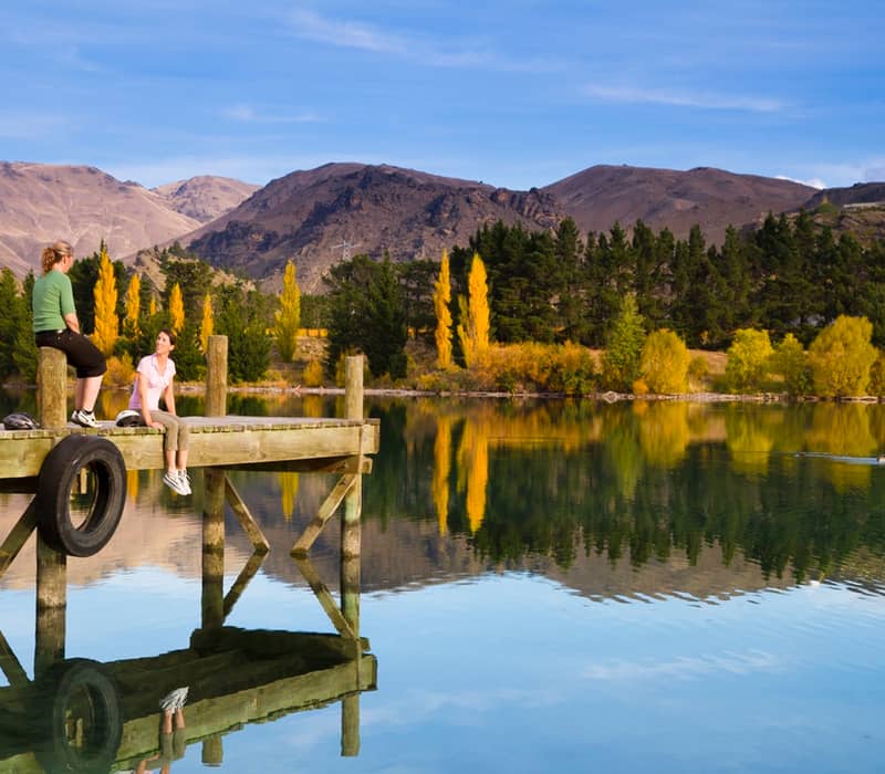 Two people sitting on a dock at a lake with autumn trees and mountains reflected in the water. Credit: Cheeky Kiwi Travel