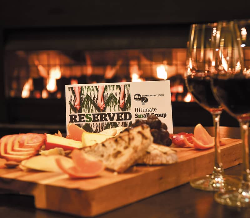 A wooden cheese platter with grapes and bread next to two glasses of red wine in front of a warm fireplace. Credit: Grand Pacific Tours