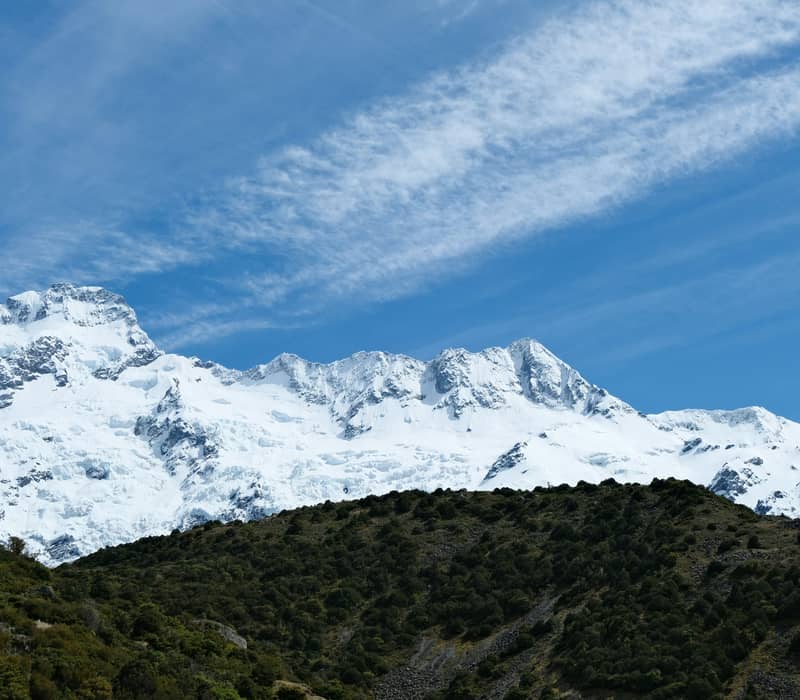 A wide view looking up past a green, scrub-covered hillside toward the massive, snow-covered peaks and hanging glaciers of Mount Sefton under a blue sky with wispy clouds. Credit: Rebecca Clarke - Unsplash