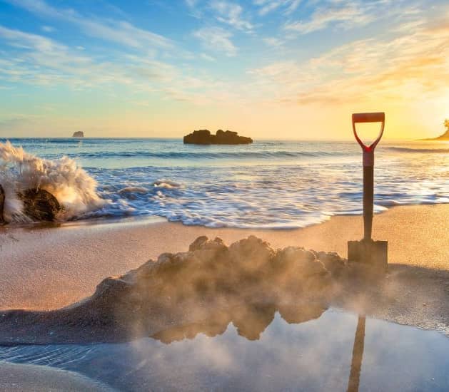Shovel in sand on beach with sunrise, waves, and rocks nearby. Credit: Pure Discoveries