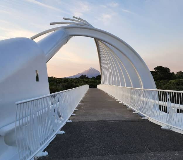 A white, modern pedestrian bridge with a skeletal, rib-like arch framing a perfectly symmetrical, snow-capped volcanic mountain in the distance under a pale sunset sky. Credit: Leisure Time Travel