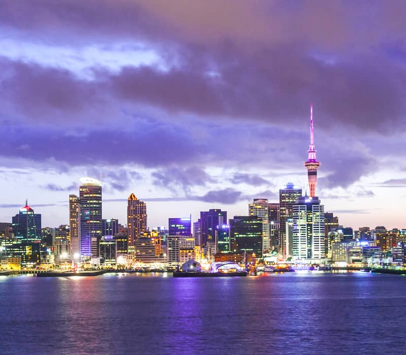 Auckland city skyline at twilight with purple clouds and city lights reflecting on the harbour water. Credit: New Zealand Fine Touring / Photographer Unknown