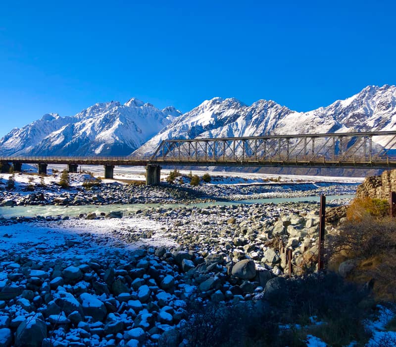 Metal bridge over a river with snow-covered rocks and mountains under a blue sky. Credit: Cheeky Kiwi - Aoraki Mt Cook