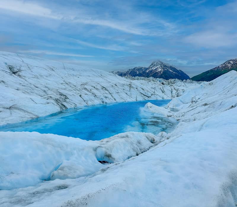 A vibrant, bright blue meltwater pool nestled within the icy crevasses and white ice ridges of a glacier. Credit: Stephen Crane - Unsplash