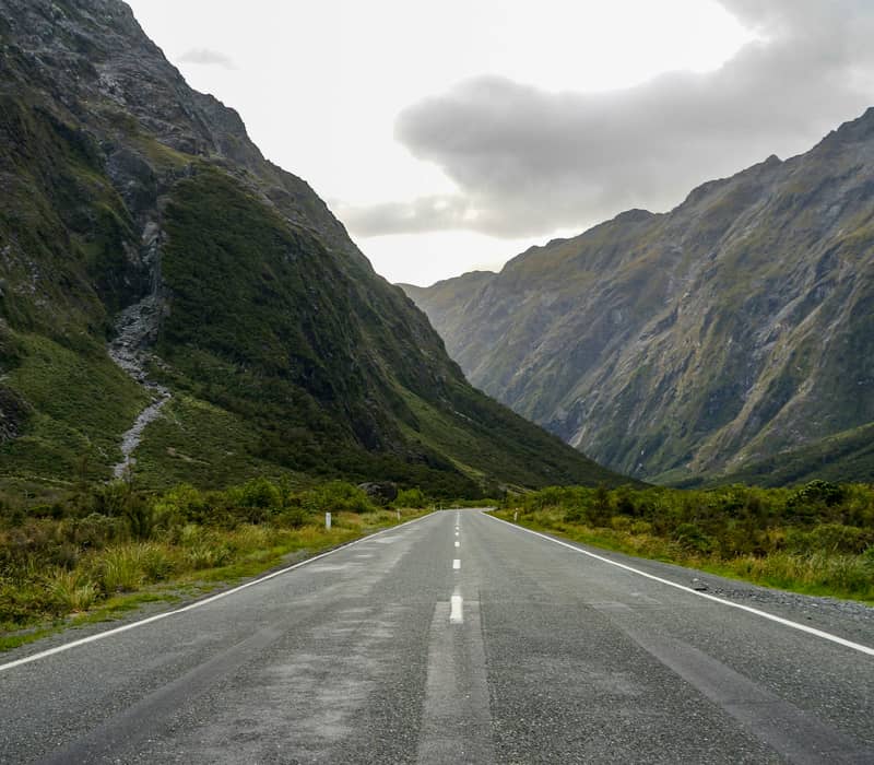 A long, straight asphalt highway stretches directly into a deep, steep alpine mountain valley flanked by massive rocky cliffs under an overcast sky. Credit: Geoff Byron - Unsplash