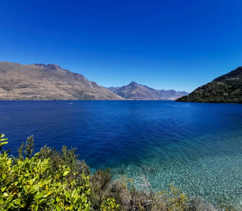 A wide-angle view across the vibrant blue and crystal-clear turquoise waters of Lake Wakatipu toward massive, rugged mountain ranges under a clear, deep blue sky. Credit: Unsplash