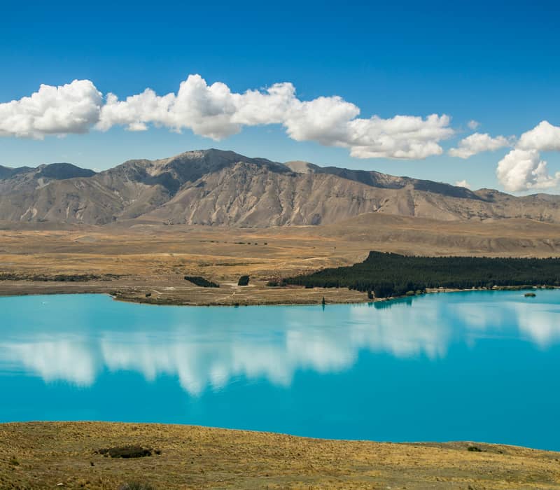 A scenic view looking down onto the vibrant turquoise waters of Lake Tekapo, showing a perfect mirror reflection of fluffy white clouds beneath dry, golden mountains. Credit: Char Beck - Unsplash