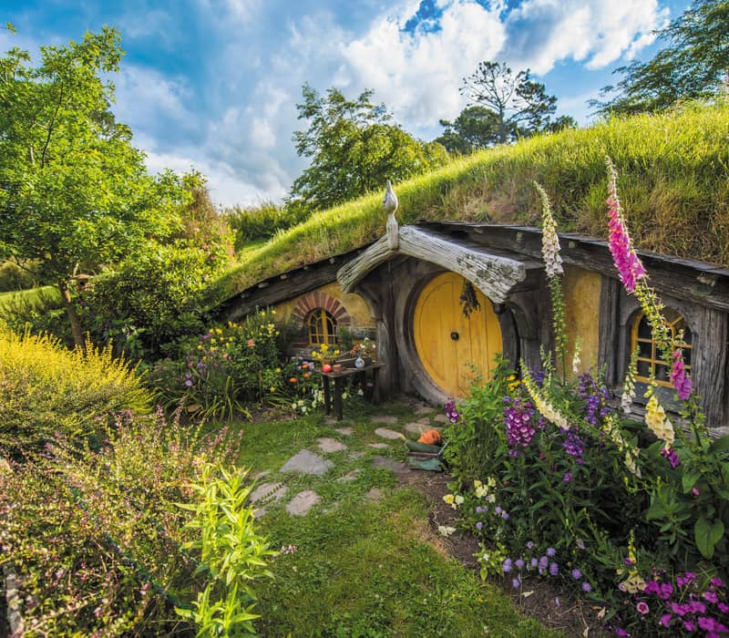 Hobbiton hobbit house entrance with round yellow door surrounded by colorful flowers and greenery under a blue sky. Credit: New Zealand Fine Touring