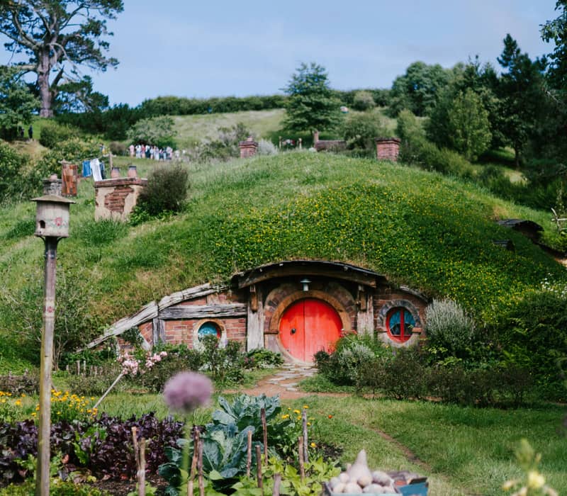 A quaint Hobbit hole with a bright round red door built into a green grassy knoll, with a detailed vegetable garden plot, wheelbarrow, and small wooden table in the foreground. Credit: Connor Gan - Unsplash