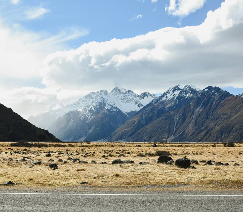 A wide roadside view looking across a rocky, golden tussock valley floor toward towering, snow-capped peaks of the Southern Alps under a partly cloudy sky. Credit: Nikhil Prasad - Unsplash