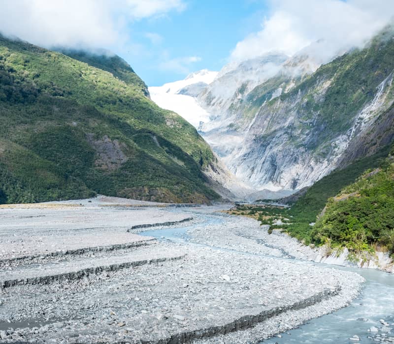 Franz Josef Glacier flowing through mountain valley with river and green forest under blue sky Credit: New Zealand Fine Touring