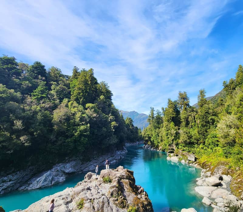 Bright blue river flowing through green forest with rocky banks and mountains under a clear sky. Credit: Hokitika Scenic Tours