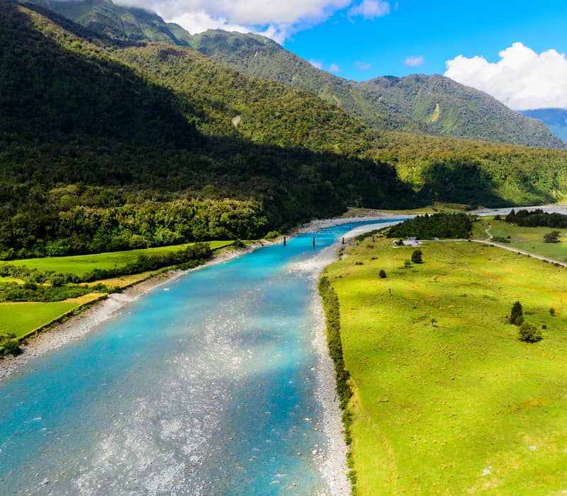 Blue river flowing through green valley with forested mountains in background under a partly cloudy sky. Credit: New Zealand Fine Touring