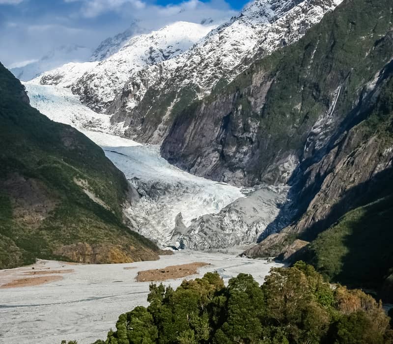 Franz Josef Glacier visible between mountains with river and wild plants in the foreground under cloudy sky. Credit: Fine Tours Group