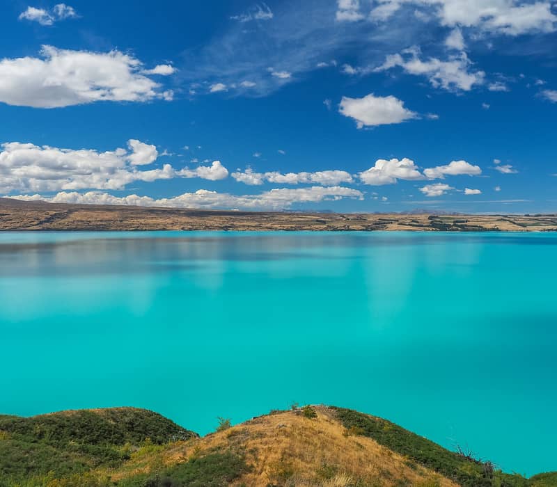 Turquoise Lake Pukaki reflecting clouds with mountains and forest on shore under blue sky