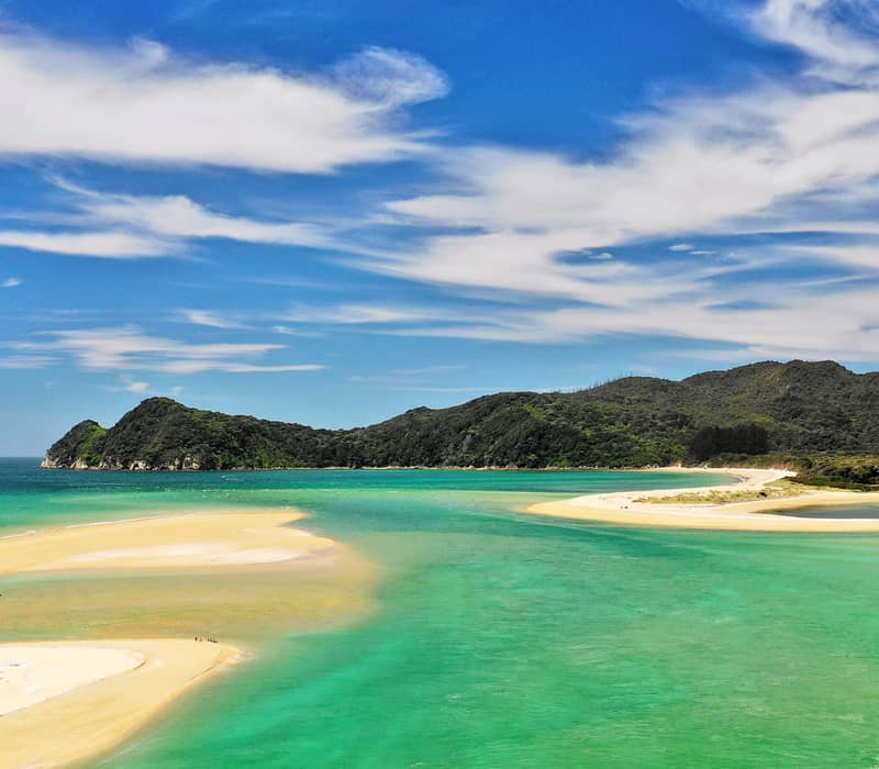 Sandy beach with turquoise water, green hills, and blue sky with clouds. Credit: Pure Discoveries