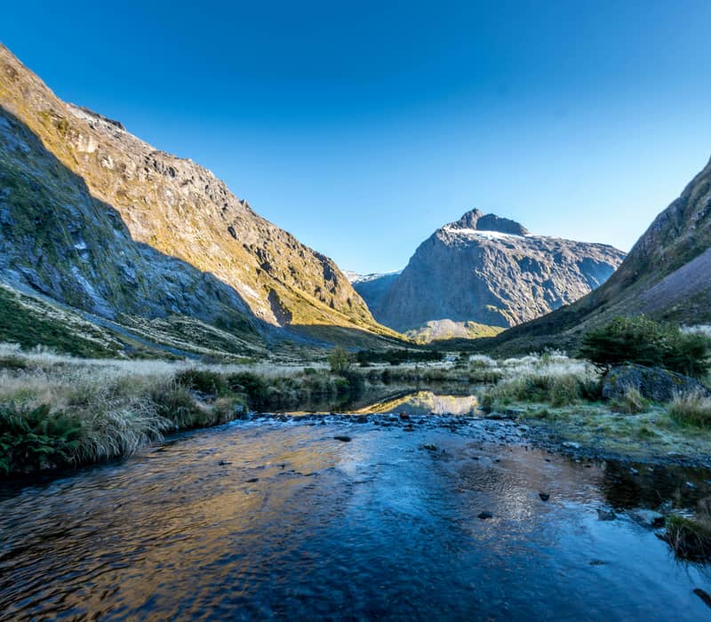 A clear glacier-fed stream flows over a rocky bed in a wide alpine valley surrounded by steep mountain cliffs under a clear blue sky. Credit: Eddie Mark Blair - Unsplash
