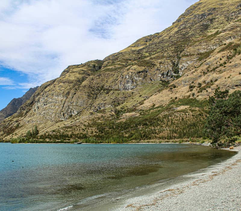 A curved pebble beach along the clear blue waters of Lake Wakatipu with the steep, tussock-covered slopes of Walter Peak rising in the background. Credit: Mikyla Eden - Unsplash