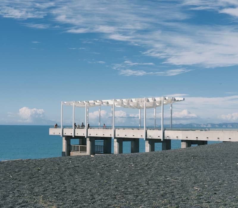 A modern concrete viewing platform with a distinct white geometric canopy extending over a dark pebble beach toward a bright blue ocean under a sunny sky. Credit: Hailey Hou - Unsplash
