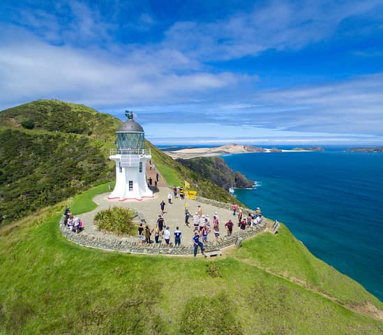 Lighthouse on a grassy hill overlooking the ocean with tourists around it on a clear day. Credit: Explore Ltd