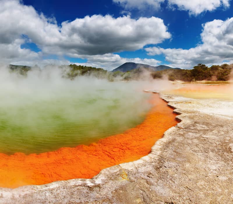 A wide geothermal pool with bright green water and a vivid orange mineral edge surrounded by thick rising steam. Credit: Grand Pacific Tours