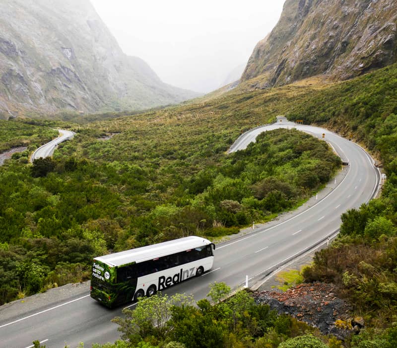 Tour bus on winding road through green valley with tall rocky mountains in the background. Credit: Real Journeys