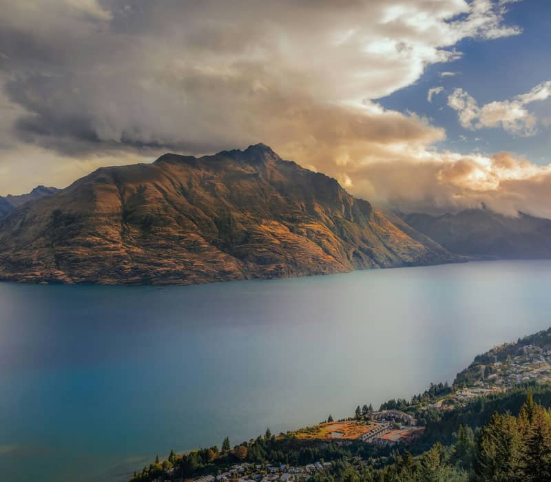 A high-angle view looking across the deep blue waters of Lake Wakatipu toward the sunlit slopes of Cecil Peak under a dramatic, cloudy sunset sky. Credit: Sam Ferrara - Unsplash