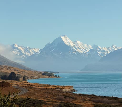 A vertical shot of a winding lakeside road leading toward the towering, snow-capped peak of Aoraki / Mount Cook across the bright blue waters of Lake Pukaki. Credit: Neve Faed