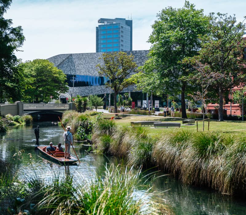 People punting on a calm river with green trees, grass, and urban buildings in Christchurch. Credit: Christchurch NZ