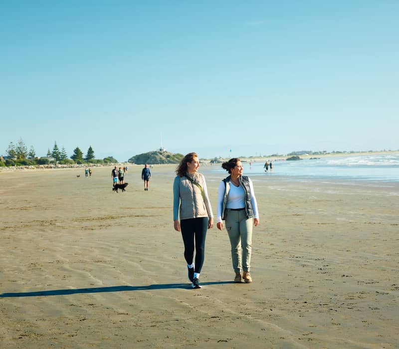 Two women walking on a sandy beach with ocean waves and people in the distance on a sunny day.