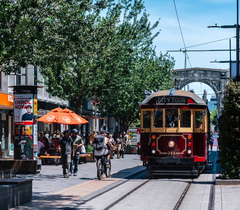 Vintage red and yellow tram on tracks with people walking and cycling in a tree-lined city mall on a bright day