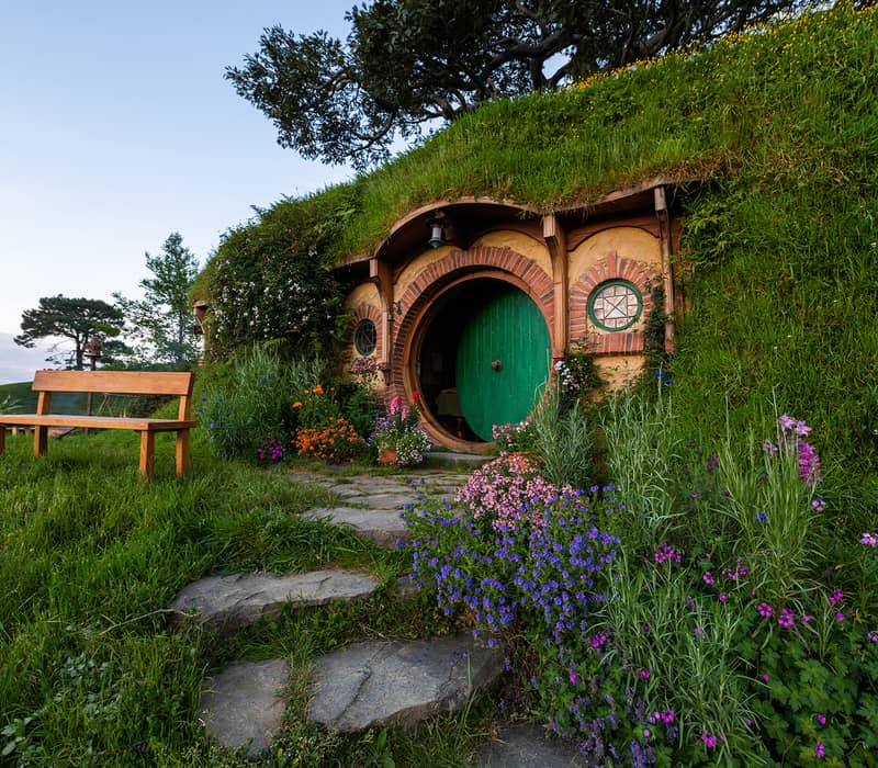 Hobbit hole with a green round door, surrounded by flowers and a wooden bench outside. Credit: Hobbiton Movie Set Tours