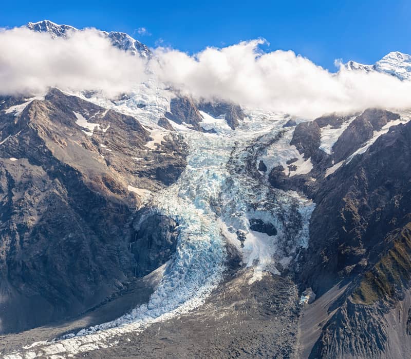 Snow-capped Fox Glacier and rocky mountains under blue sky with white clouds. Credit: Fine Tours Group