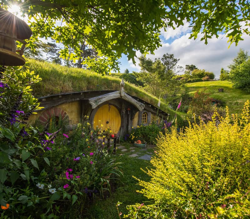 Hobbit hole with yellow door surrounded by vibrant garden and greenery under sunny sky. Credit: Hobbiton Movie Set Tours