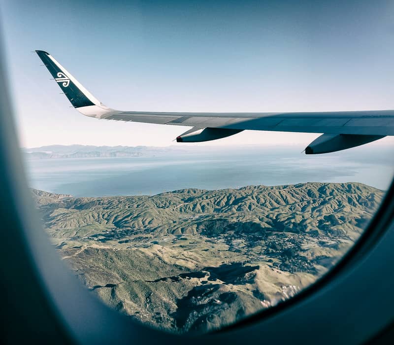 View from an airplane window of an airplane wing flying over green hills with wind turbines and the ocean. Credit: Photo by James Coleman on Unsplash
