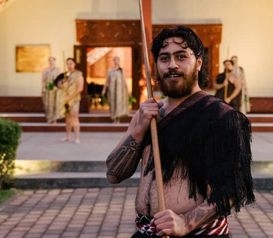 Maori warrior in traditional attire with wooden spear in front of a meeting house.